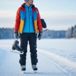 Osoba v zimním oblečení s bruslemi na rameni stojí u zamrzlého jezera na zasněžené cestě.