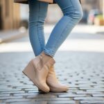Stylová žena v béžových klínových botách a modrých džínách stojí na dlážděné ulici.